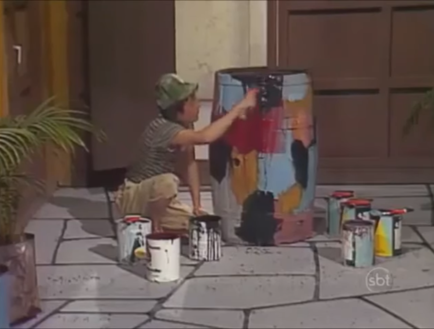 Chaves sentado no chão da vila, cercado de latas de tinta, pintando o barril com várias cores.