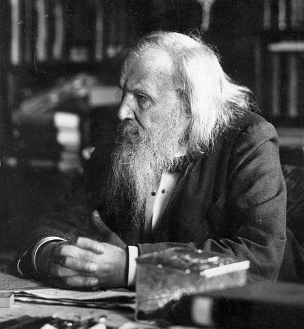 Dmitri Mendeleev, um homem com cabelos médios e barba longa, sentado. A foto está em preto e branco.